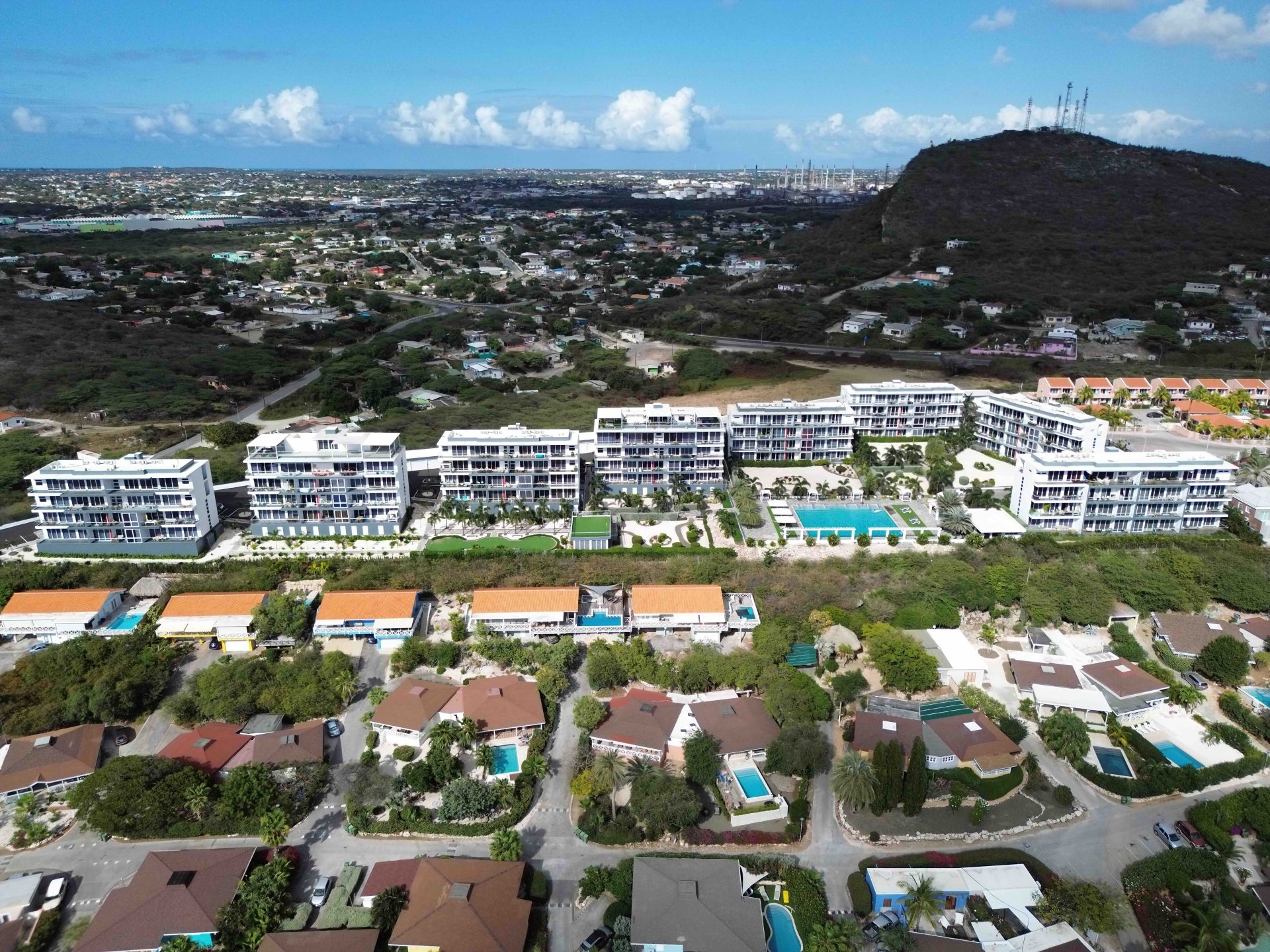 Aerial view of Grand View Residences featuring modern apartments with pools, surrounded by residential homes and lush greenery, set against a scenic landscape in Aruba.