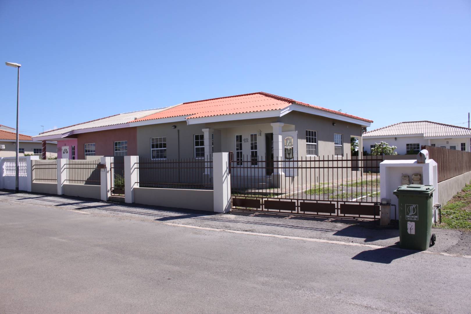 Residential property in Curaçao with a gated entrance, featuring a beige exterior and red-tiled roof, surrounded by a well-maintained yard and nearby homes.