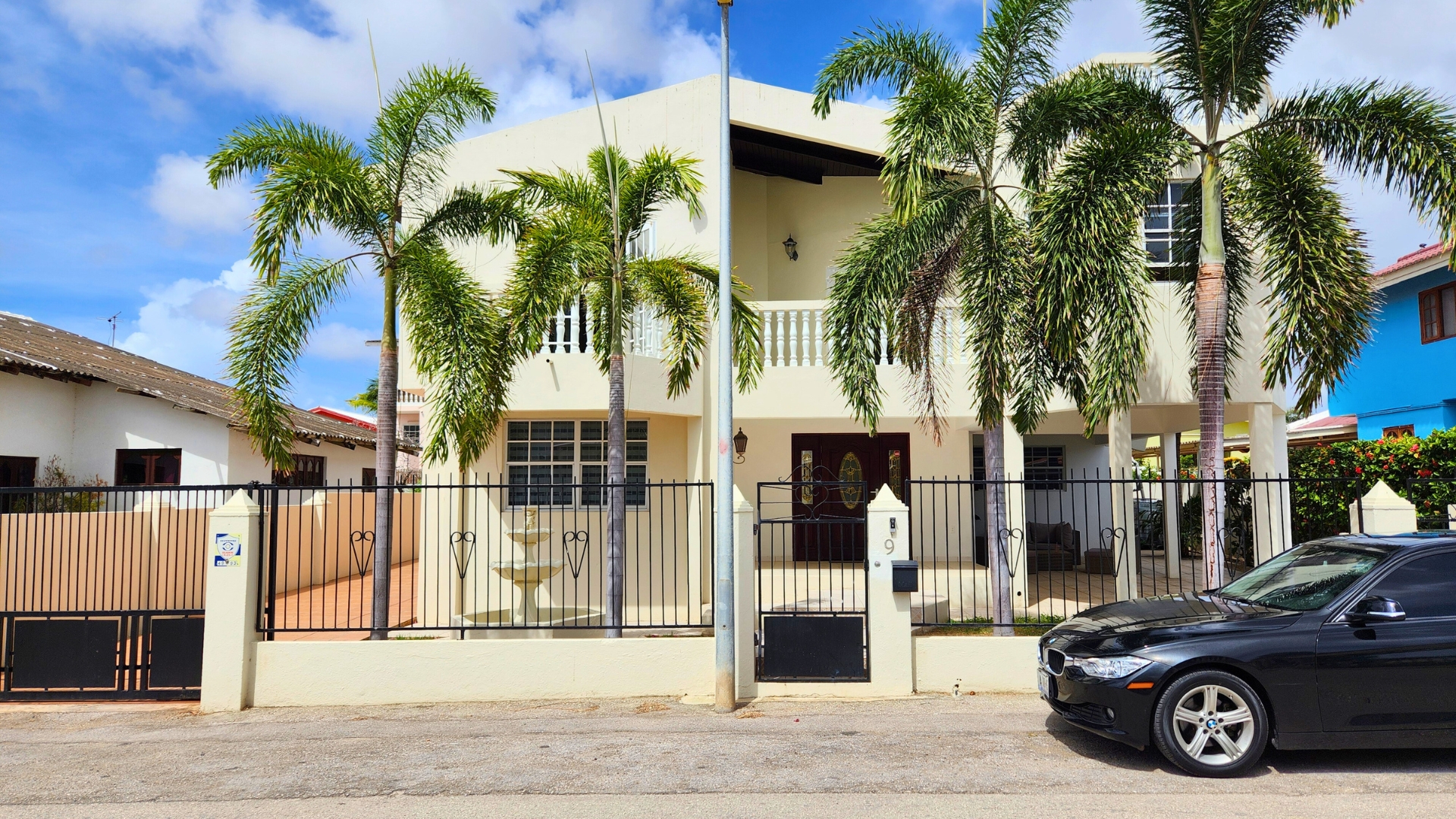 Modern two-story house with palm trees, black car parked in front, and decorative iron fence, showcasing a welcoming entrance.