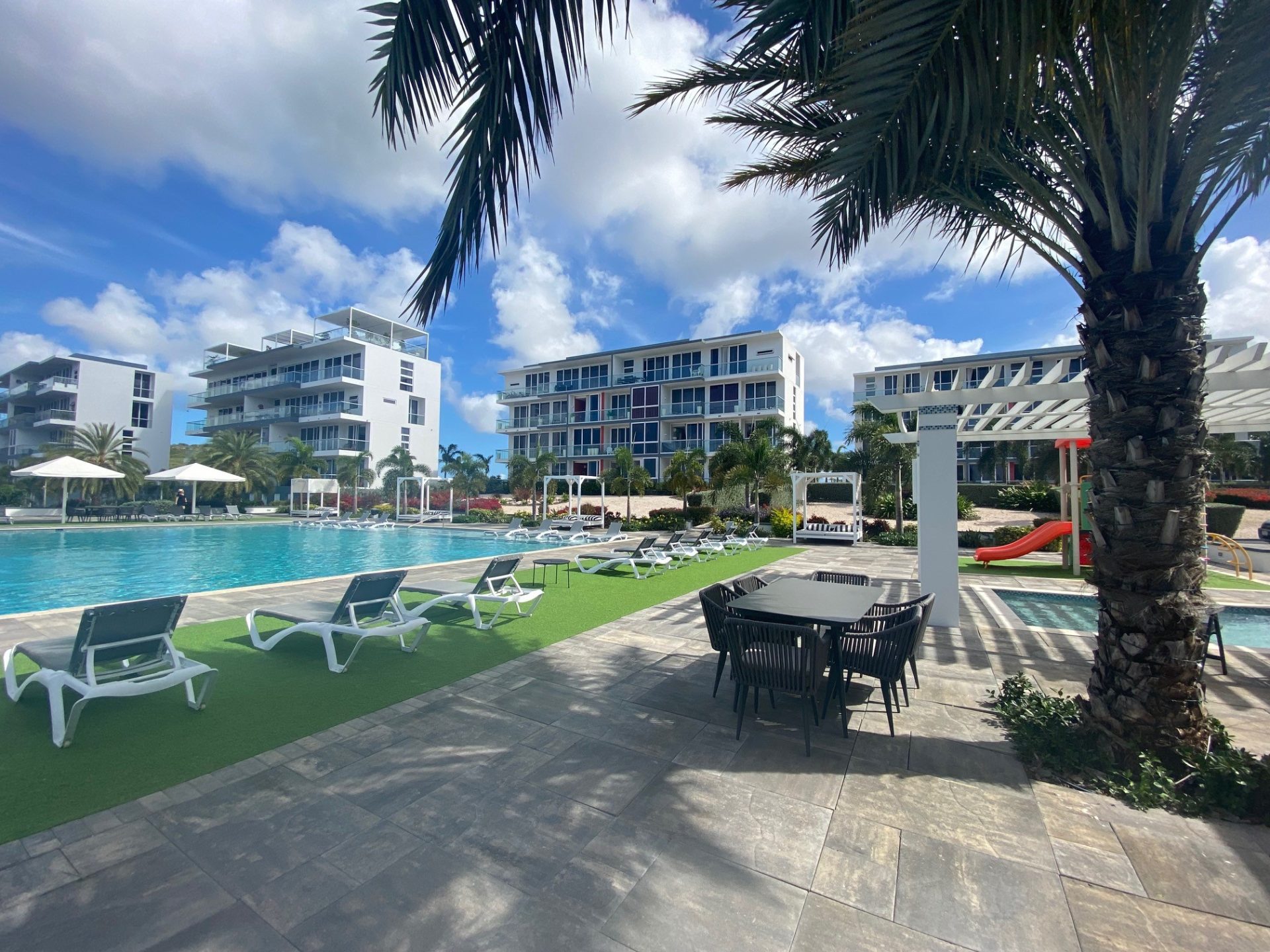 Modern resort-style pool area with lounge chairs, palm trees, and residential buildings under a blue sky.