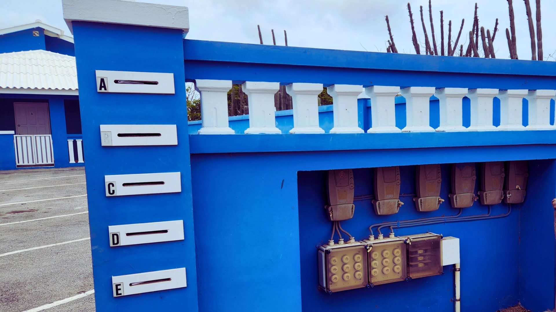 Blauwe muur met brievenbussen gemarkeerd met letters A, C, D en E, en elektriciteitsmeters, voor een huurwoning in Abrahamsz, Curaçao.