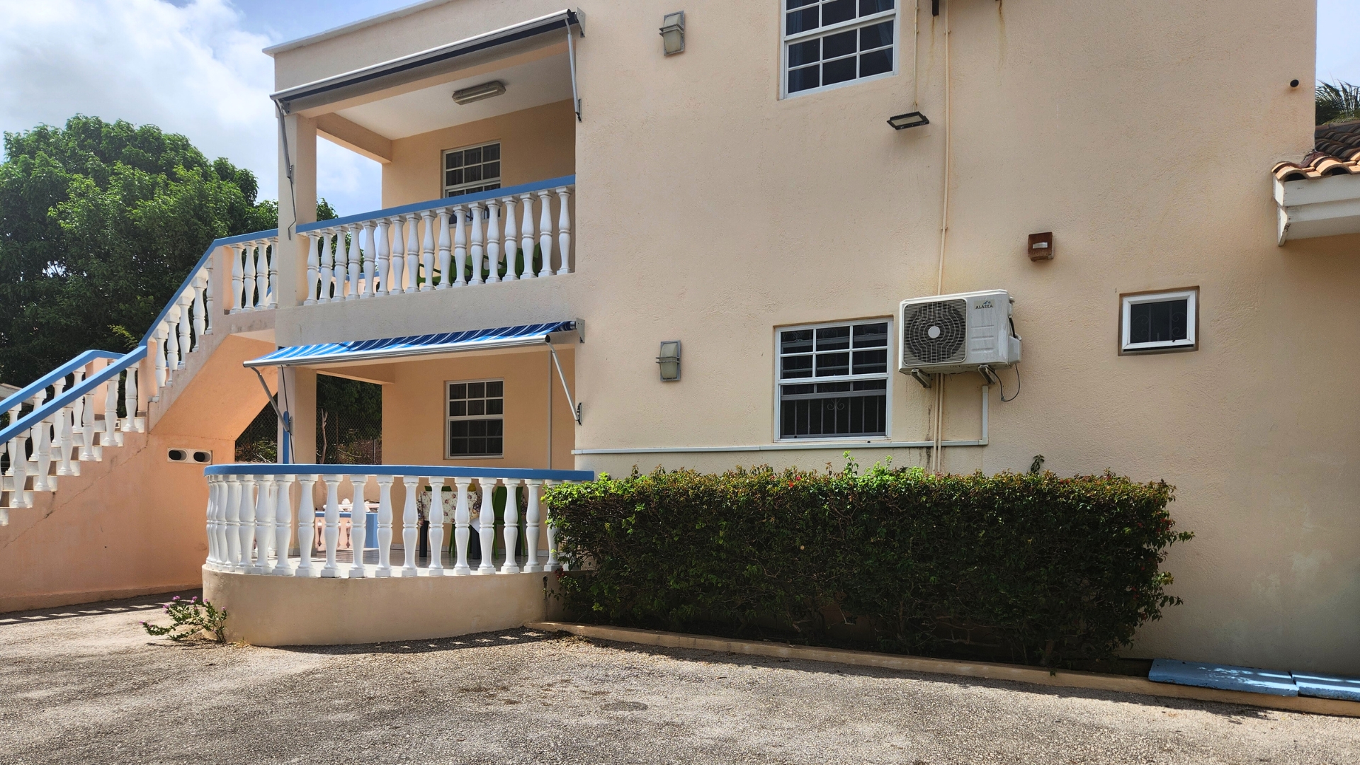 Exterior view of a two-story building with a staircase, white railings, air conditioning unit, and green shrubbery, showcasing a sunny day.