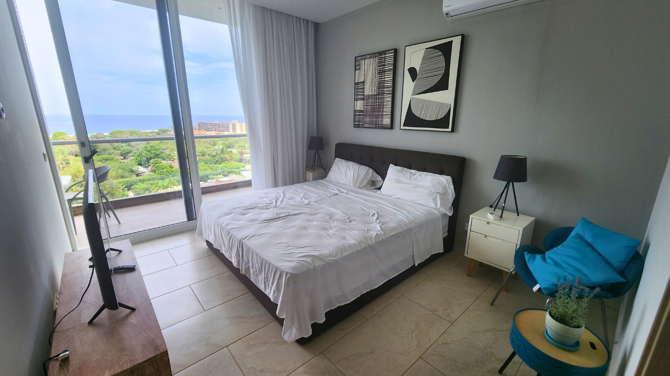 Modern bedroom with a queen-sized bed, white linens, decorative wall art, a TV stand, and a view of greenery and ocean through large glass doors.