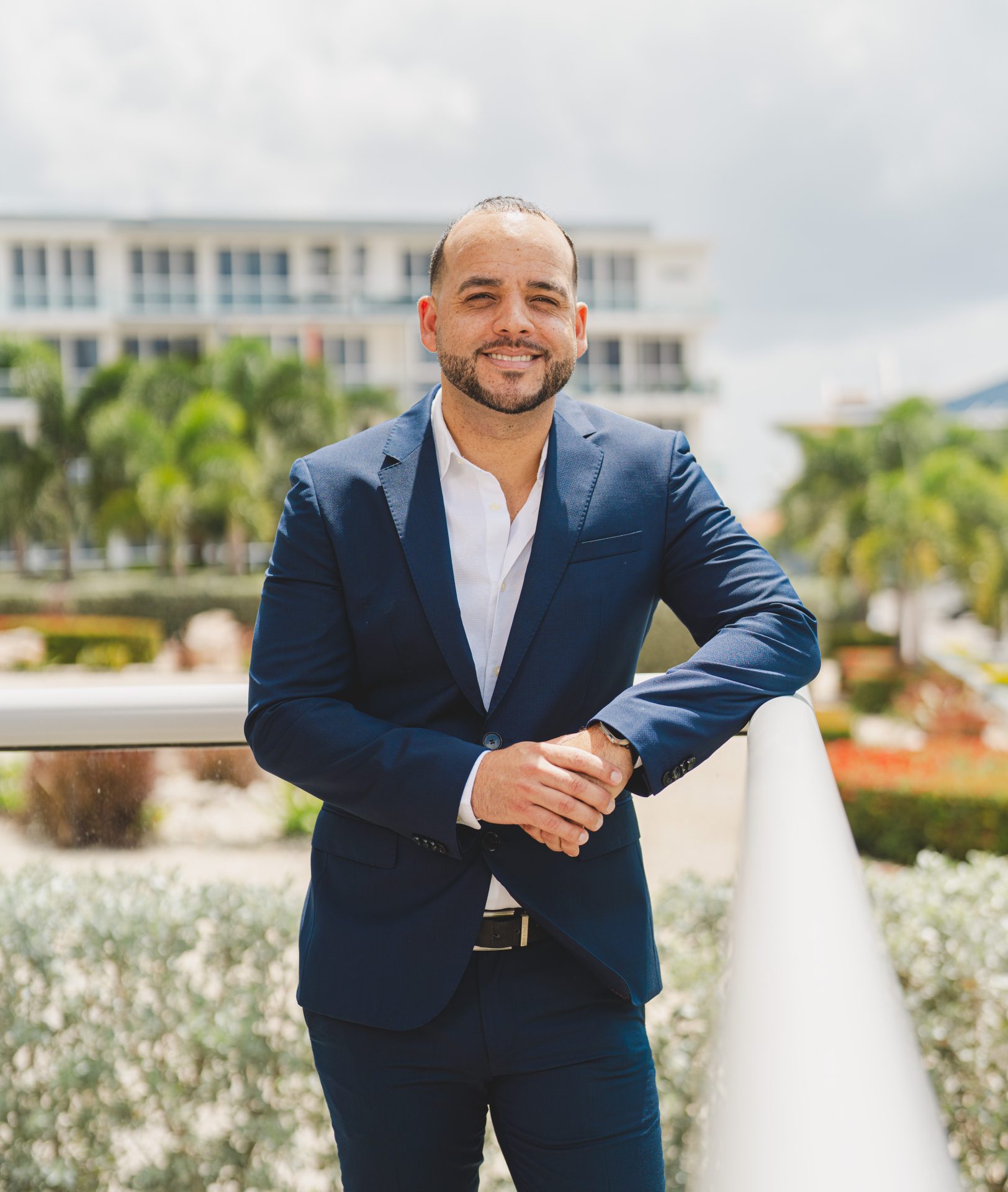 Man in a blue suit smiling confidently, standing outdoors near a railing with lush greenery and modern buildings in the background, representing New Winds Realty's professional image in luxury real estate.