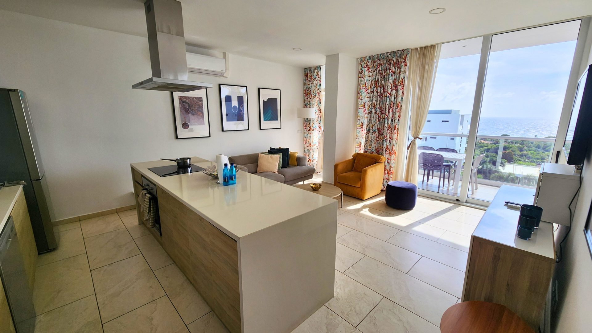 Luxurious penthouse interior featuring modern kitchen, stylish living area with sofa and armchair, vibrant curtains, and ocean view from large windows.