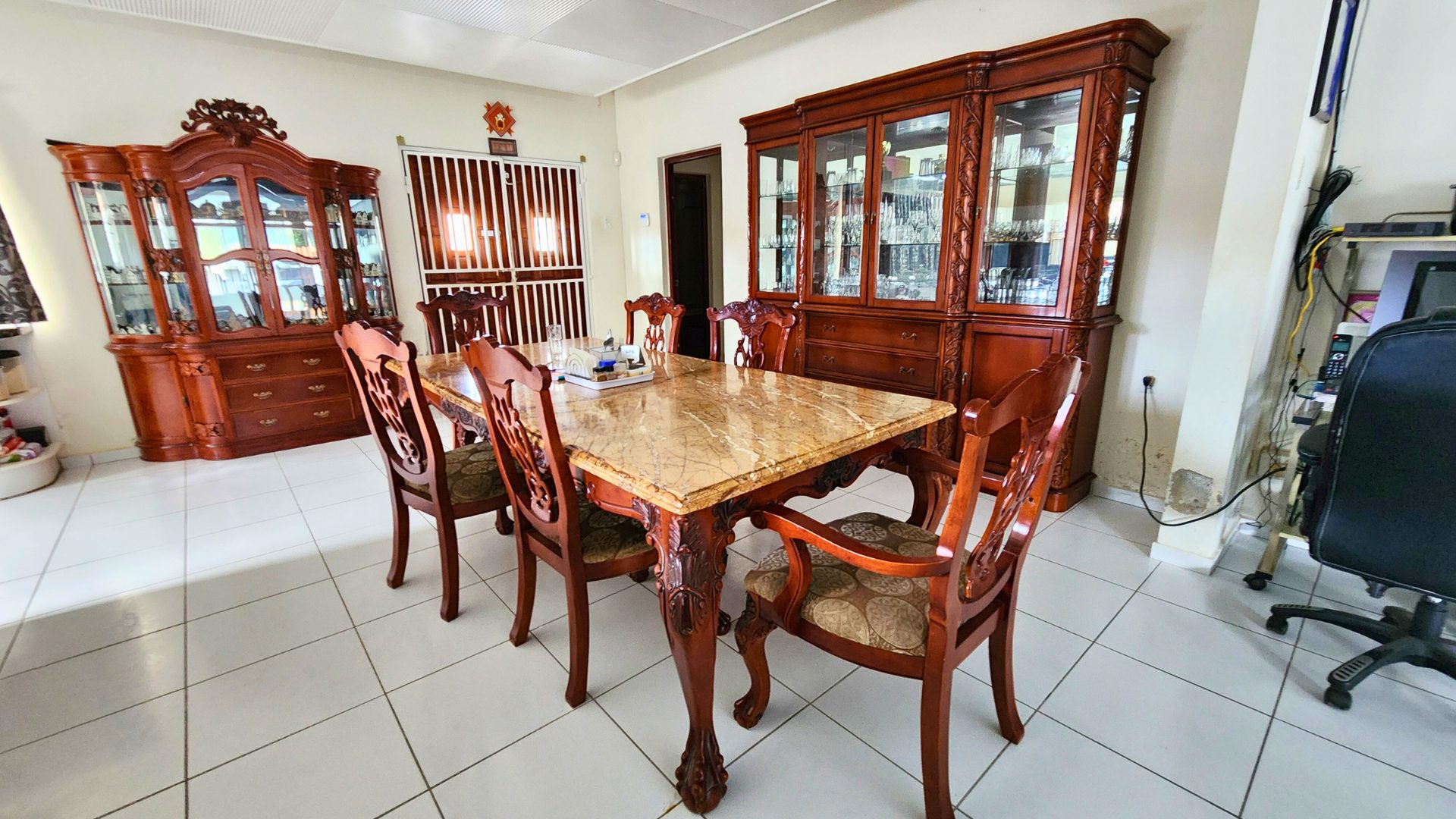 Dining room featuring a wooden table with marble top, surrounded by ornate chairs, and a glass-fronted cabinet displaying collectibles, emphasizing family-friendly living in a four-bedroom property for sale by New Winds Realty.