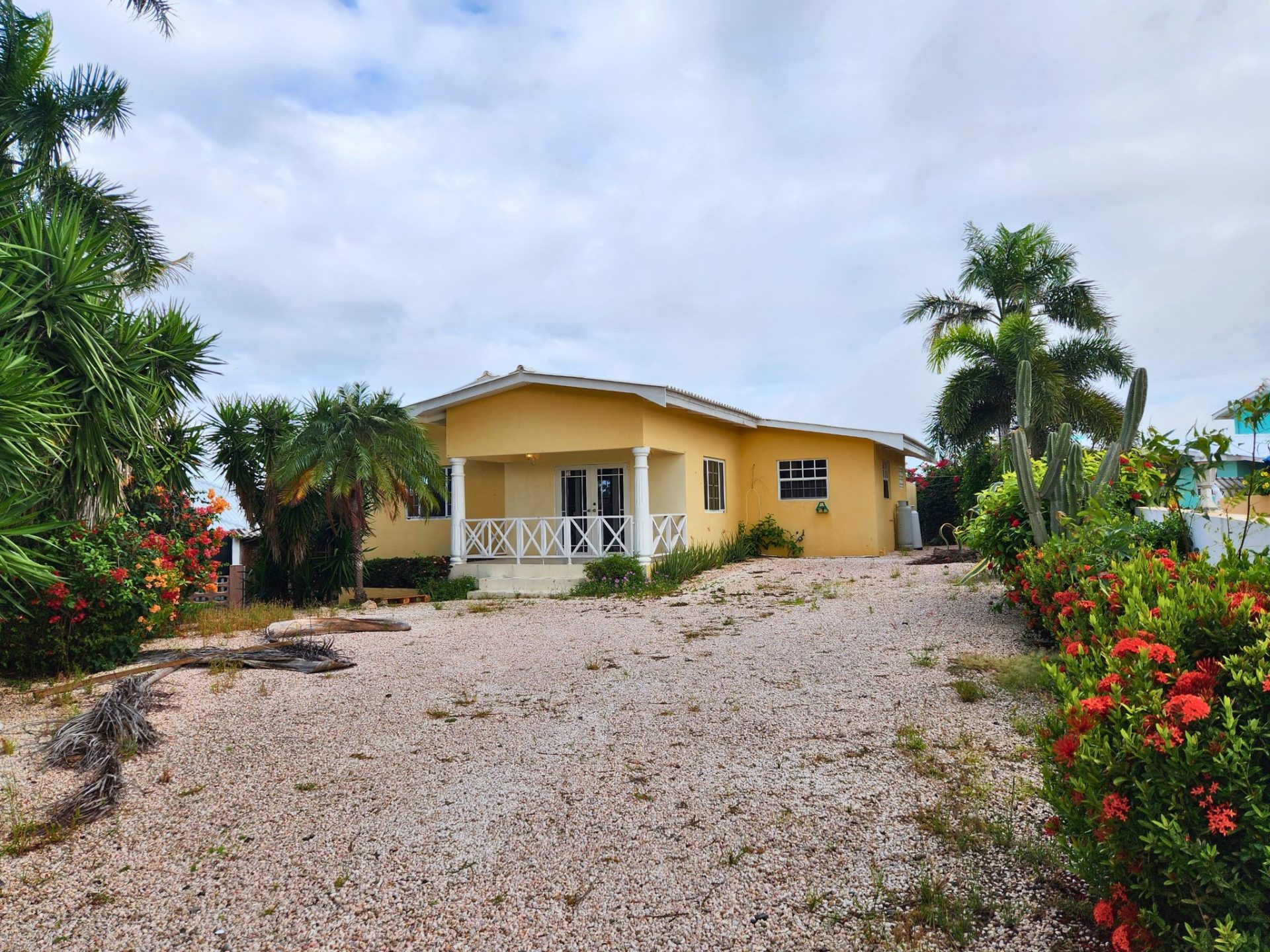 Yellow house surrounded by tropical plants and flowers, gravel driveway, and cloudy sky.