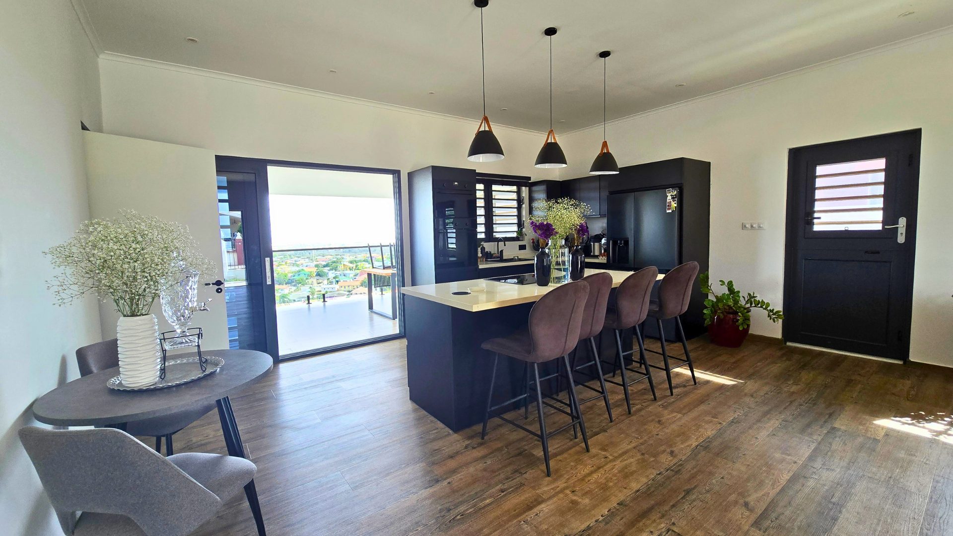 Moderne keuken met eiland, barstoelen, en uitzicht op de omgeving, inclusief bloemen in vazen en een deur naar het balkon in het huis 'Wonen met Uitzicht - #1606' in Sunset Heights, Curaçao.