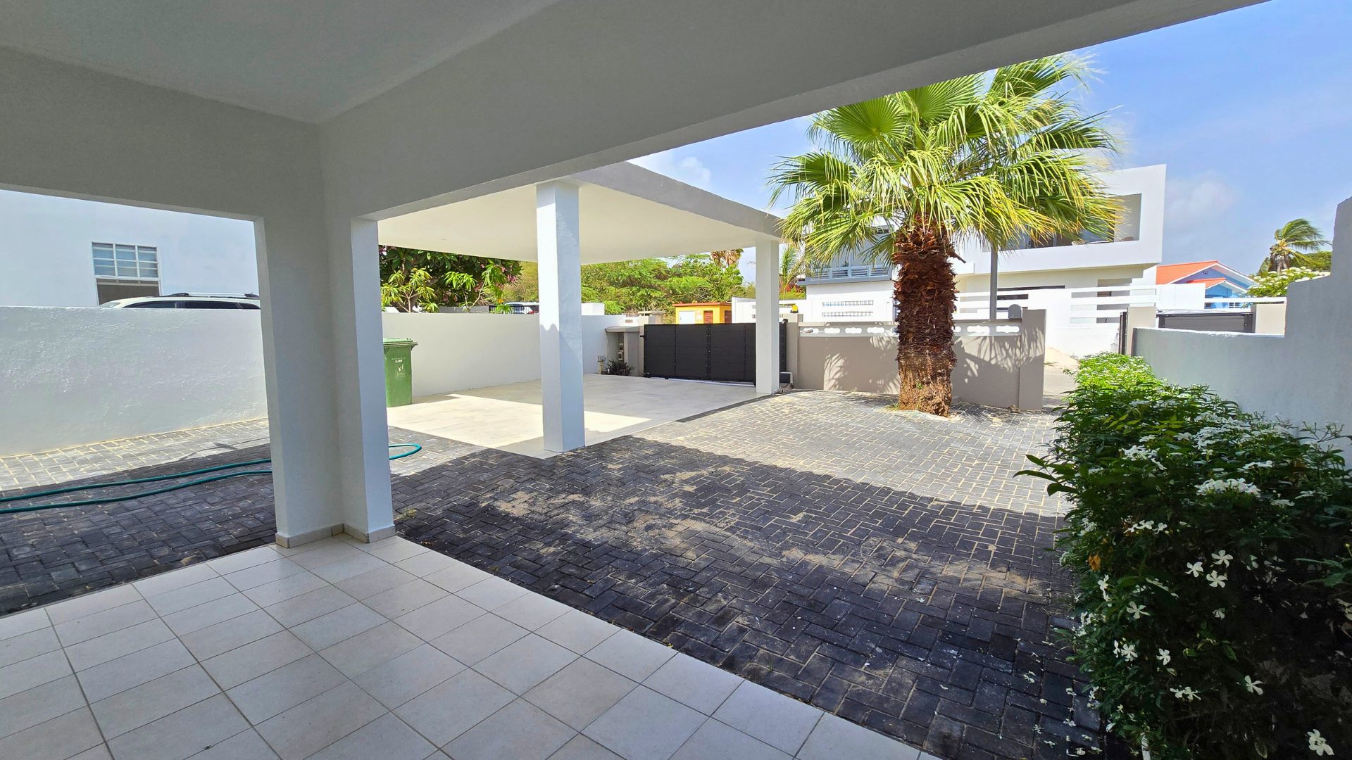 Veranda view of a rental property in Sunset Heights, Curaçao, featuring palm trees, paved driveway, and a clean, modern exterior.