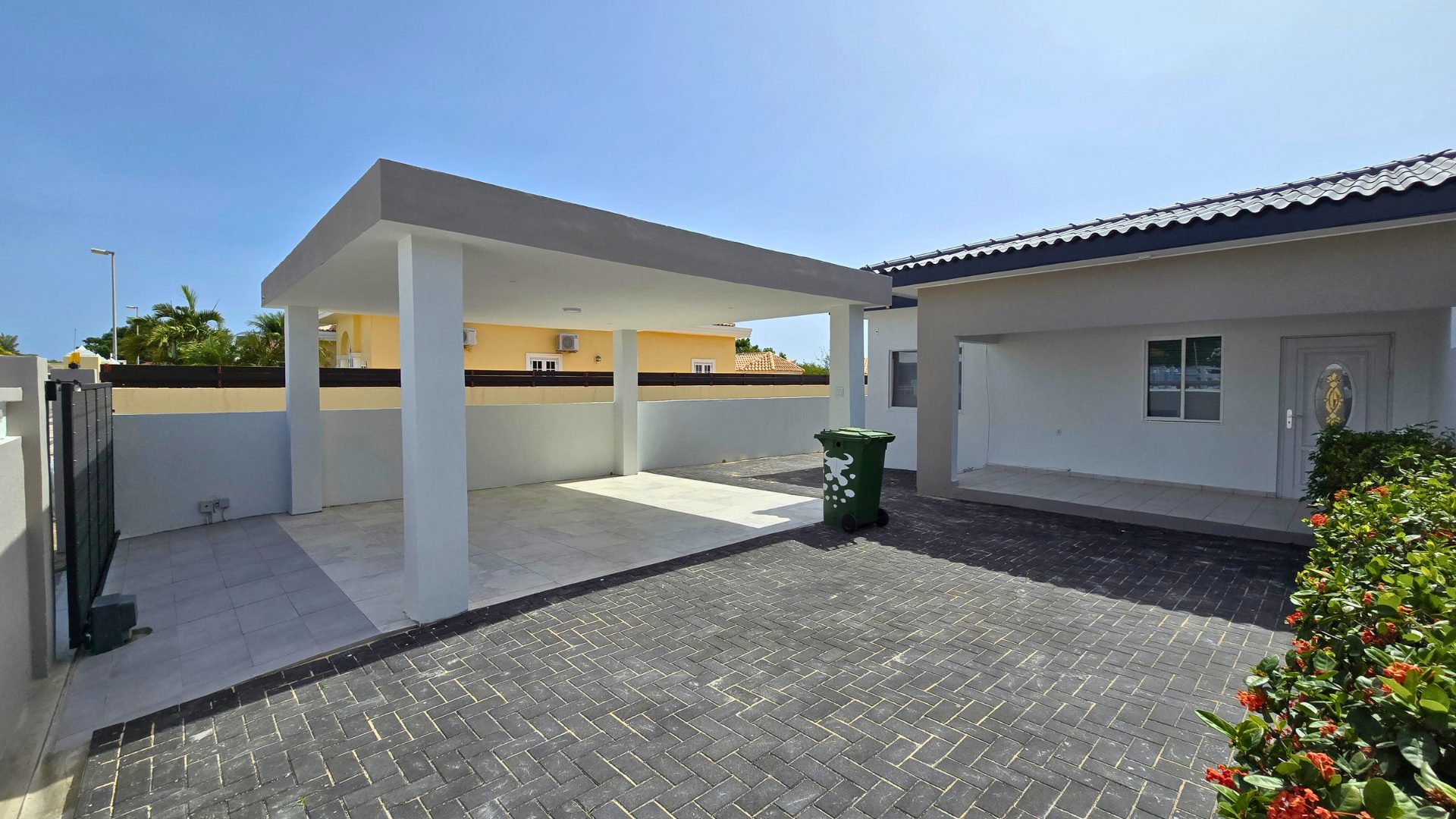 Veranda and entrance of a rental home in Sunset Heights, Curaçao, featuring a paved driveway, green trash bin, and surrounding greenery.